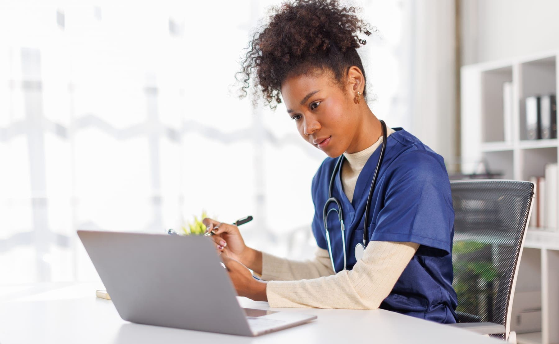 A nurse in blue scrubs writing while working at a laptop.