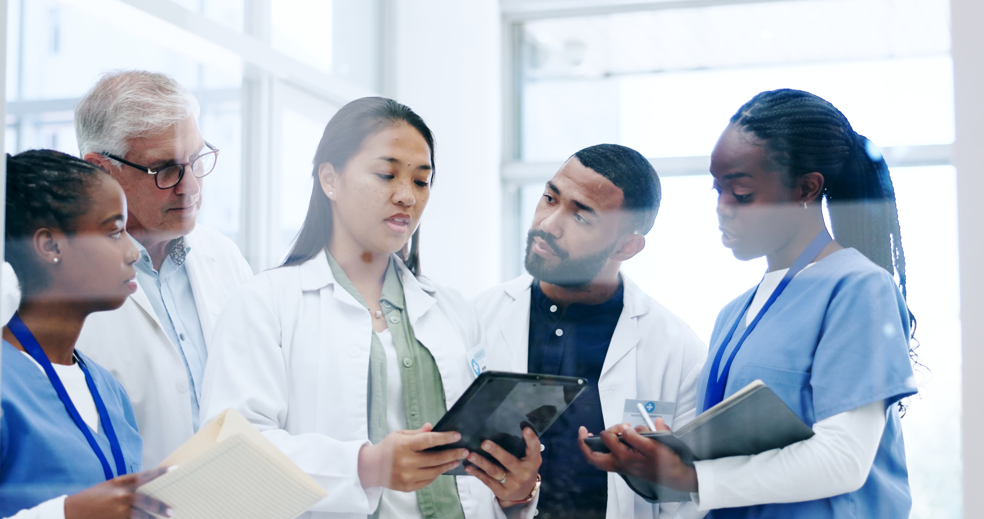 A diverse clinical team gathered around a tablet in a bright hospital setting, reviewing information together.